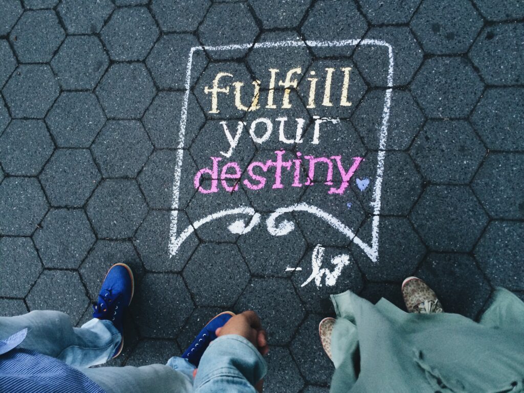 A chalk-written message on a hexagonal paved ground, reading “fulfill your destiny” in colorful letters. Two individuals are partially visible.