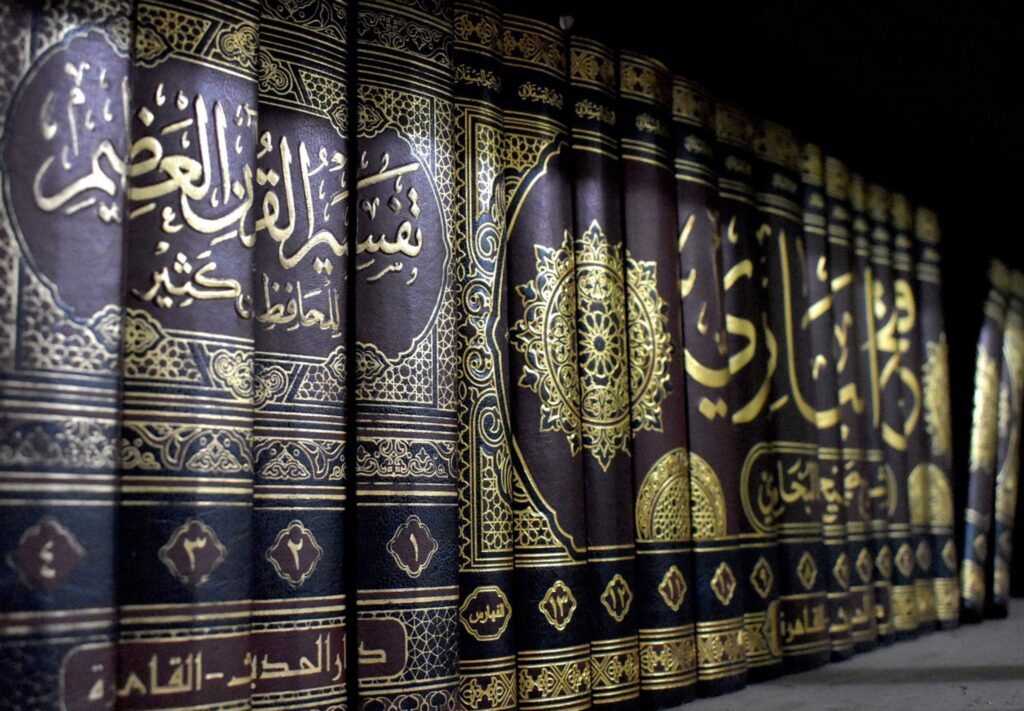 A bookshelf containing books of Tafseer and Hadith with Arabic calligraphy and intricate golden designs on the spine.