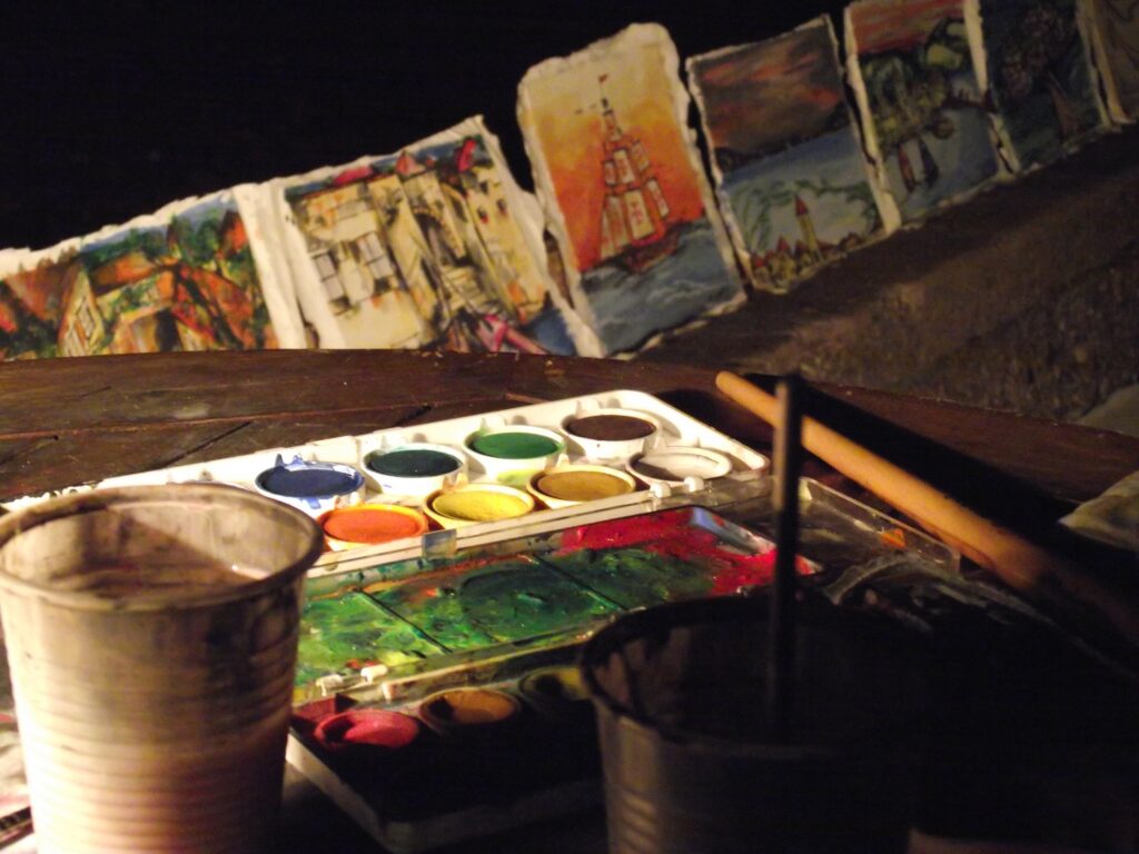 An artist’s workspace at night with watercolors, brushes, and small paintings depicting various scenes.