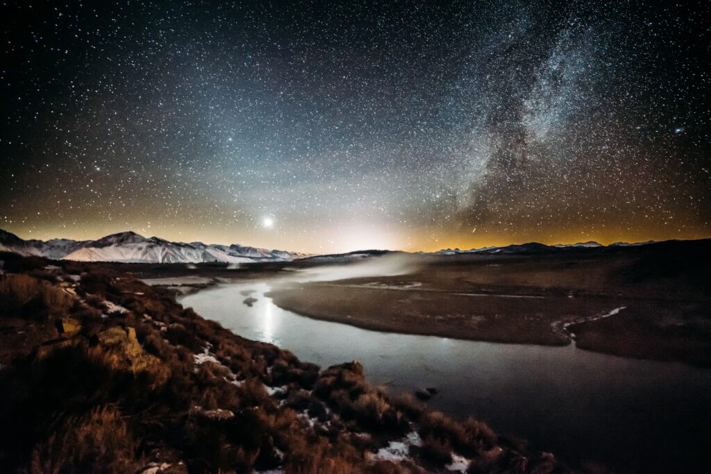 Night landscape featuring a star-studded sky, the Milky Way, and a glowing horizon illuminating a tranquil river winding through snow-capped mountains.